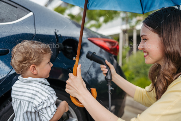 Close up of smiling mother and her little son charging their electric car on a rainy day, standing