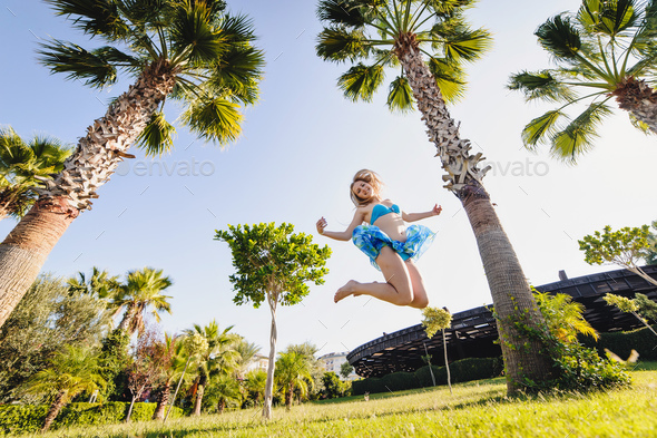 Beautiful happy young slim girl jumps between Tall palm trees. In the blue swimsuit, dress, shawl