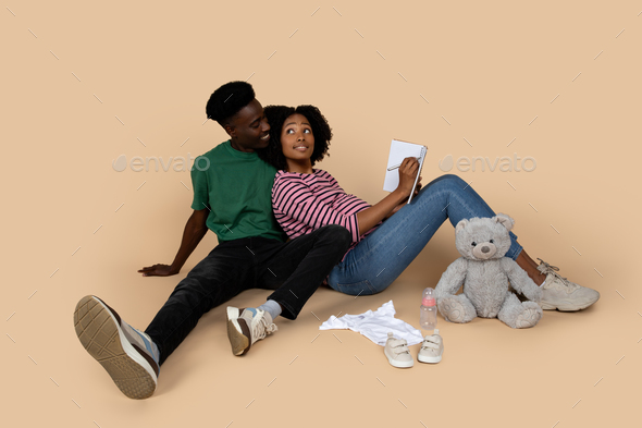 Smiling millennial black couple with baby things make notes plan, enjoy pregnancy, sit on floor