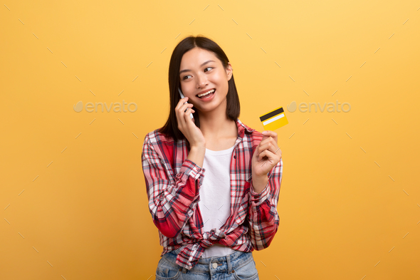 E-commerce concept. Excited asian lady holding credit card and talking on phone, speaking with
