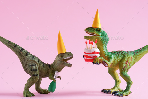 Cute plastic toys green dinosaurs in party hats with cake and champagne bottle on pink background.