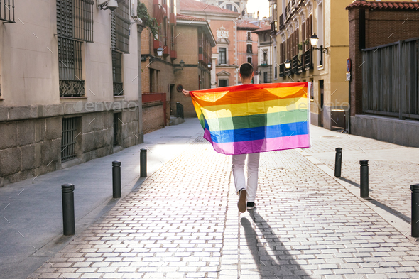 Proudly Embracing the LGBT Flag, a Backlit Man Stands with Open Arms on Sunny City Street at Sunset