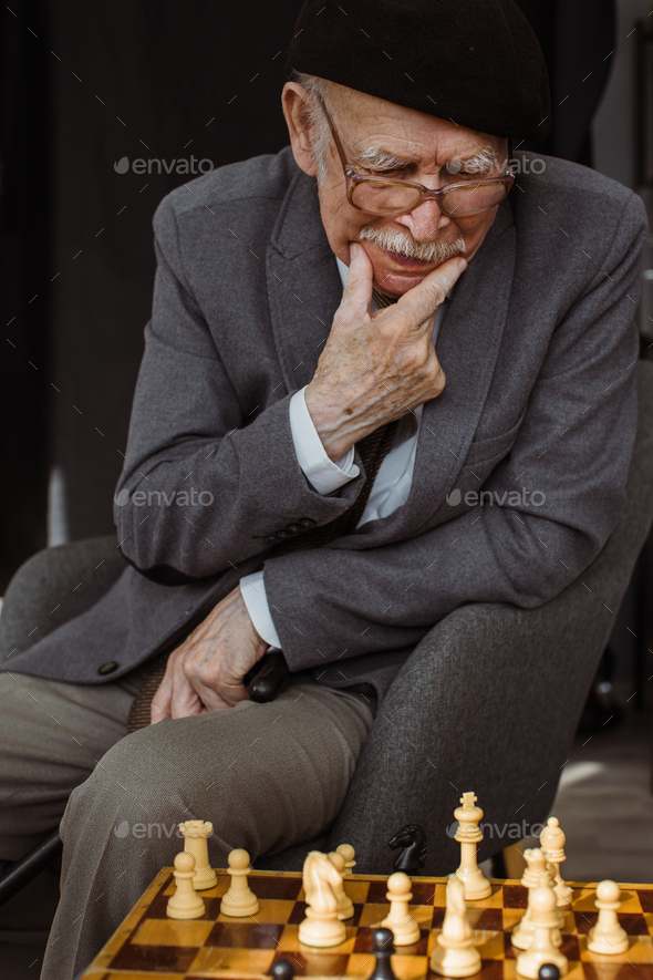 Old man in checked shirt holding glasses and thinking about next chess move  in game with imaginary opponent Stock Photo