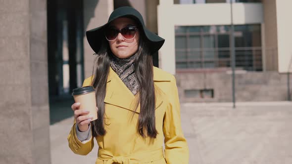Stylish Young Girl in a Yellow Raincoat Hat and Glasses Walks Down the Street Looking Around alt