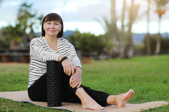 Smiling middle aged sportive woman in casual sportswear sitting on yoga mat and relaxation after