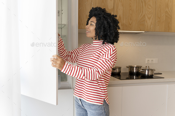 Beautiful, attractive African American woman opening refrigerator in stylish modern kitchen