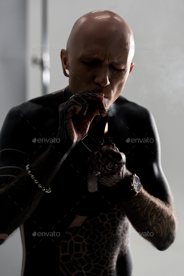 bare-chested man with tattoos lighting cigar with lighter on grey