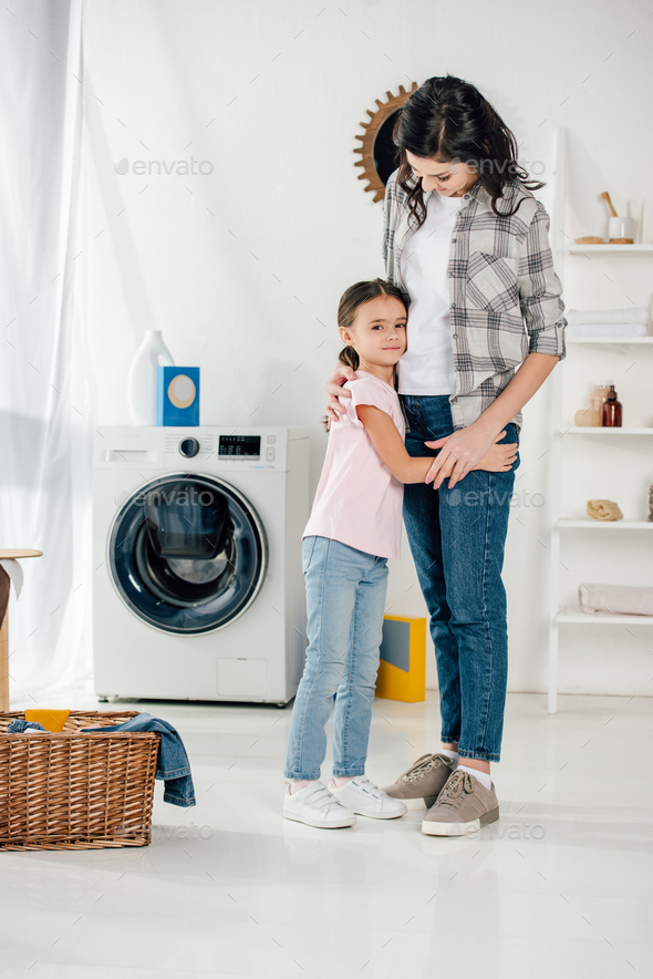 daughter in pink t-shirt hugging smiling mother in grey shirt in laundry room