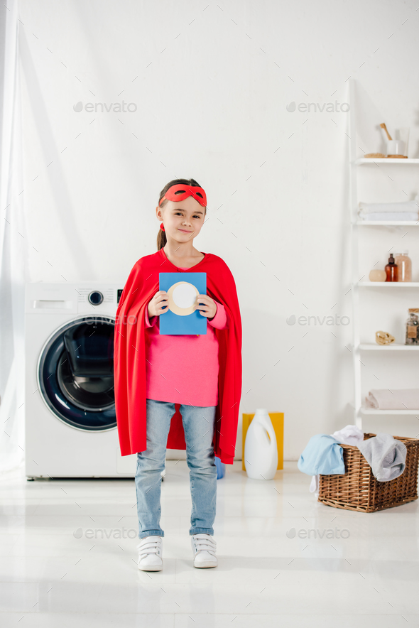 child standing in red homemade superhero suit and holding washing