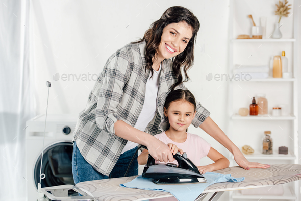 smiling mother in grey shirt and daughter in pink t-shirt ironing in laundry room