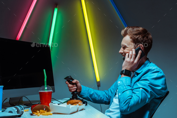 side view of smiling and handsome cyber sportsman holding joystick and talking on smartphone