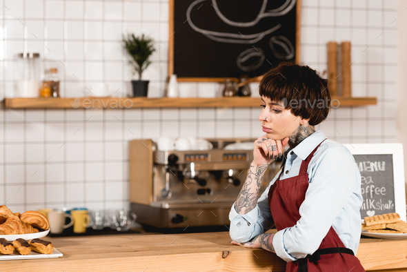 pensive barista in apron standing near wooden bar counter in coffee shop