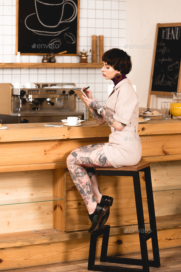 fashionable, tattooed businesswoman sitting at bar counter and writing in notebook