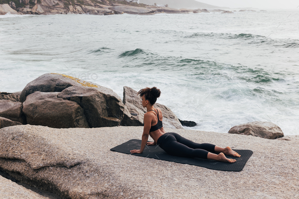 Female in cobra yoga pose at seaside. Woman in sportswear doing stretching  workout on mat by ocean. Stock Photo by ArtemVarnitsin