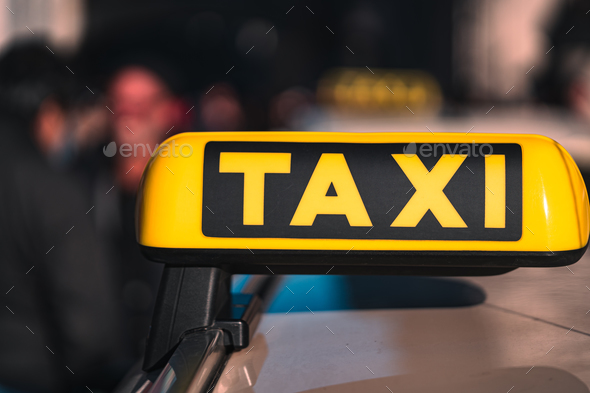 Shallow focus of a yellow TAXI sign on the top of a Taxi car with blurred people in the background