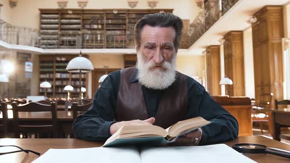 Old Bearded Man which Flipping Through Book Pages Sitting at the Library Table alt