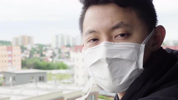 A Young Asian Man in a Face Mask Looks at the Camera As He Stands on a Balcony in an Urban Area alt
