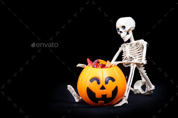 A seated skeleton pulls out of a bucket in the shape of a pumpkin candy. Trick or Treat