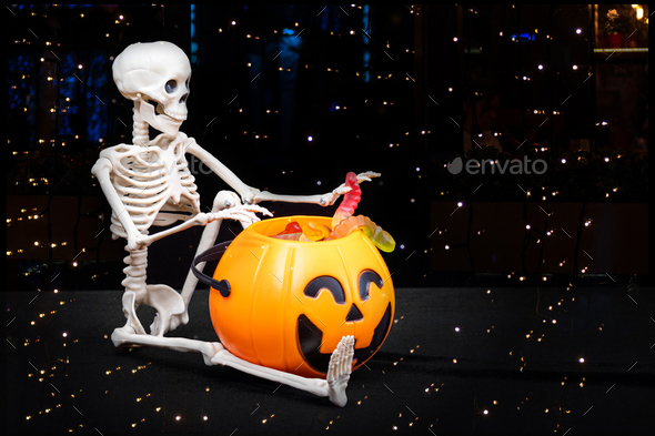 A seated skeleton pulls out of a bucket in the shape of a pumpkin candy on a black with starry bokeh