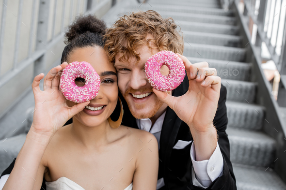 having fun, wedding in city, excited interracial newlyweds obscuring face with donuts