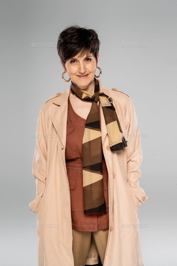 happy mature woman looking at camera on grey, fall fashion, trench coat, scarf, hoop earrings