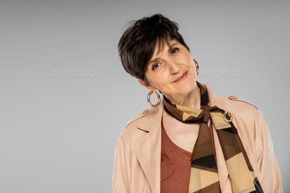 autumn fashion, brunette mature woman smiling at camera on grey, trench coat, scarf, hoop earrings