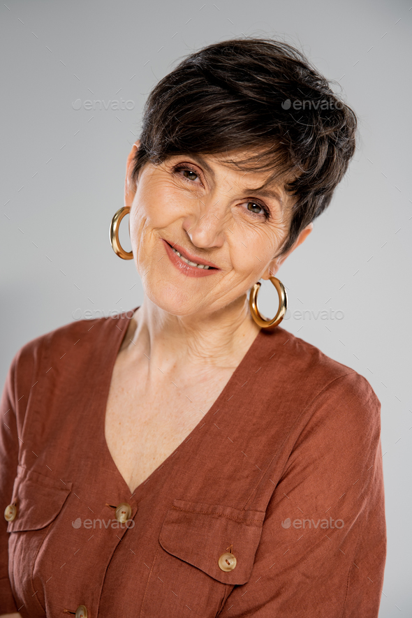 brunette middle aged woman in brown jacket and hoop earrings smiling at camera on grey, fall fashion