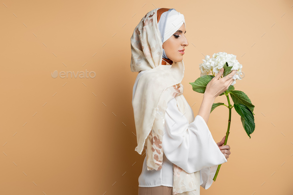 graceful muslim woman in silk headscarf and blouse holding white flower on beige, side view
