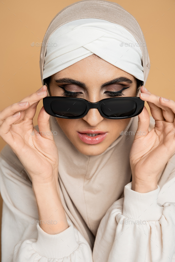 charming muslim woman in hijab taking off trendy sunglasses on beige, personal style, individuality