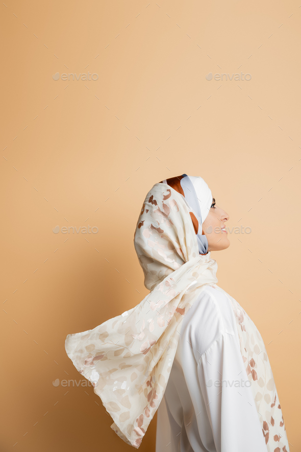 side view of smiling muslim woman in elegant silk headscarf and white blouse on beige