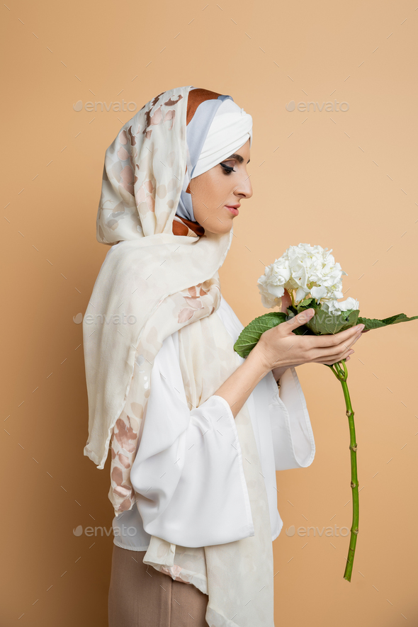 side view of trendy muslim woman in headscarf and white blouse holding hydrangea flower on beige
