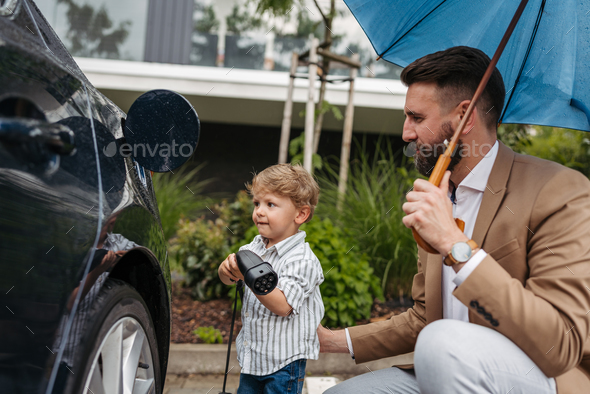 Close up of father and his little son charging their electric car on a rainy day, standing under