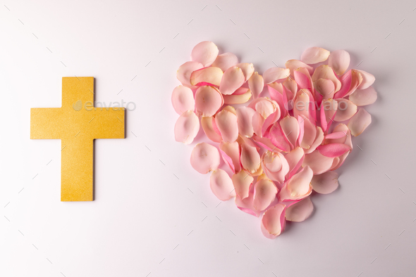 Overhead of yellow christian cross and heart shape of pink rose petals on white background