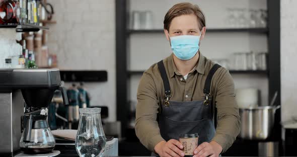 Portrait of Caucasian Handsome Male Barista in Medical Mask and Gloves Giving Cup of Coffee to alt