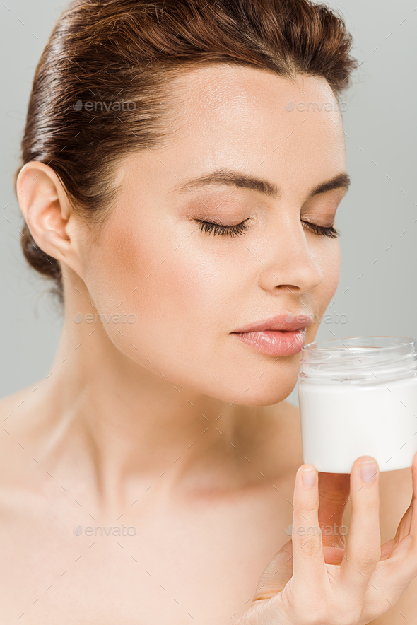 attractive woman smelling face cream and holding container isolated on grey