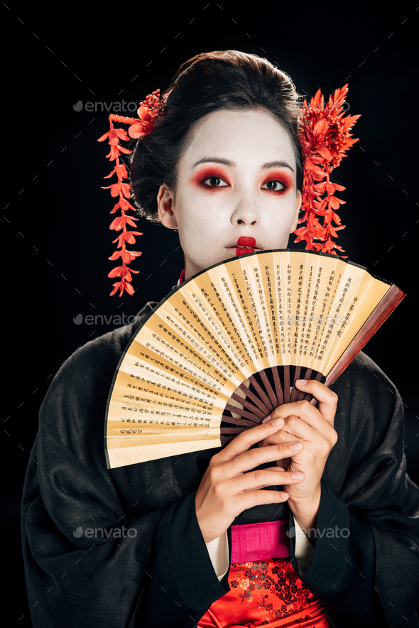 young geisha in black kimono with red flowers in hair holding traditional asian hand fan isolated on