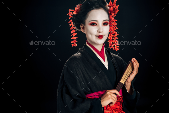 happy geisha in black kimono with red flowers in hair holding traditional asian hand fan isolated on