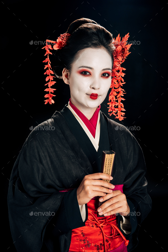 smiling geisha in black kimono with red flowers in hair holding traditional asian hand fan isolated