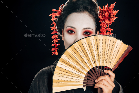geisha in black kimono with red flowers in hair holding traditional asian hand fan near face