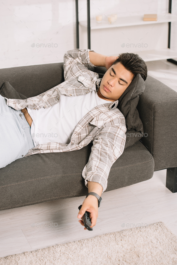 exhausted asian man sleeping on couch while holding tv remote controller
