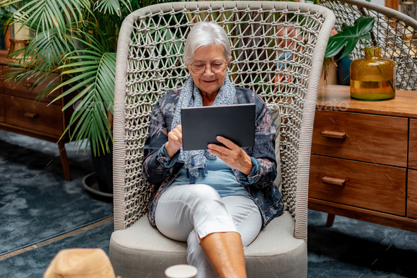 Senior white-haired woman sitting indoor using digital tablet surfing enjoying tech and social