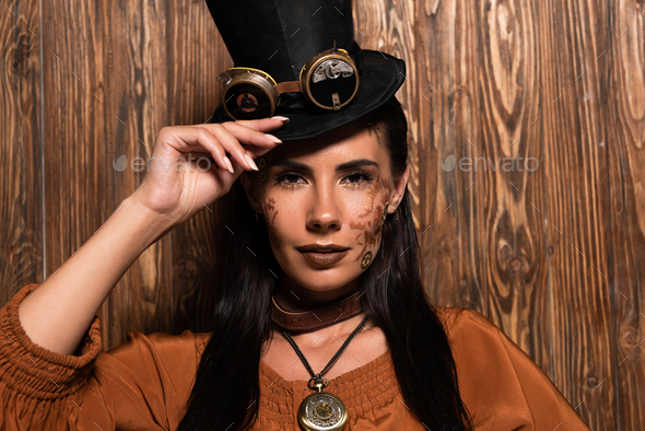 front view of steampunk woman touching top hat with goggles looking at camera on wooden