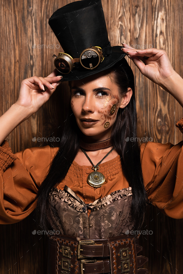 attractive steampunk woman touching top hat with goggles on wooden