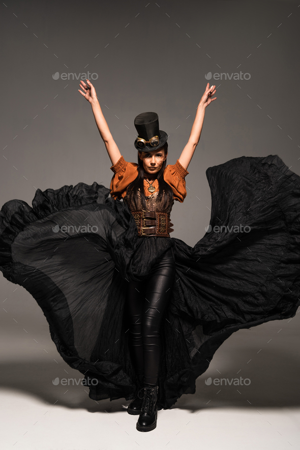 full length view of attractive steampunk woman in top hat with goggles standing with hands up on