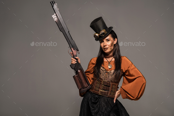 attractive steampunk woman in top hat holding gun and standing with hand on hip isolated on grey