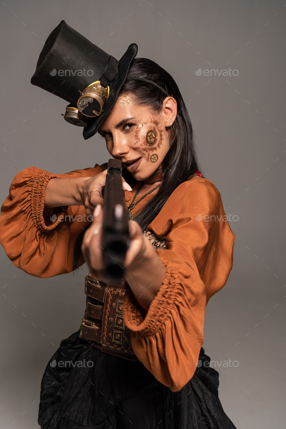 focused steampunk woman in top hat aiming with gun isolated on grey