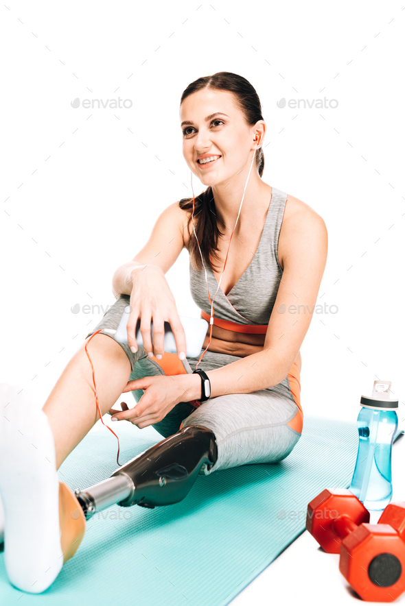 disabled sportswoman sitting on fitness mat and listening music in earphones isolated on white