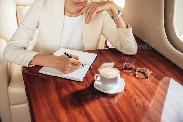 cropped view of businesswoman in suit writing in notebook in private plane