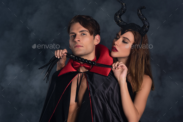 attractive woman with horns holding flogging whip near man in vampire  halloween costume on black Stock Photo by LightFieldStudios