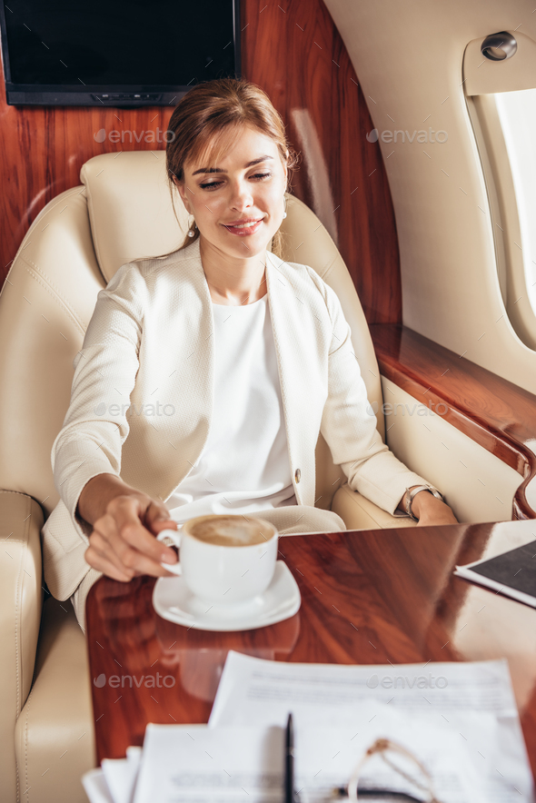smiling businesswoman in suit taking cup of coffee in private plane